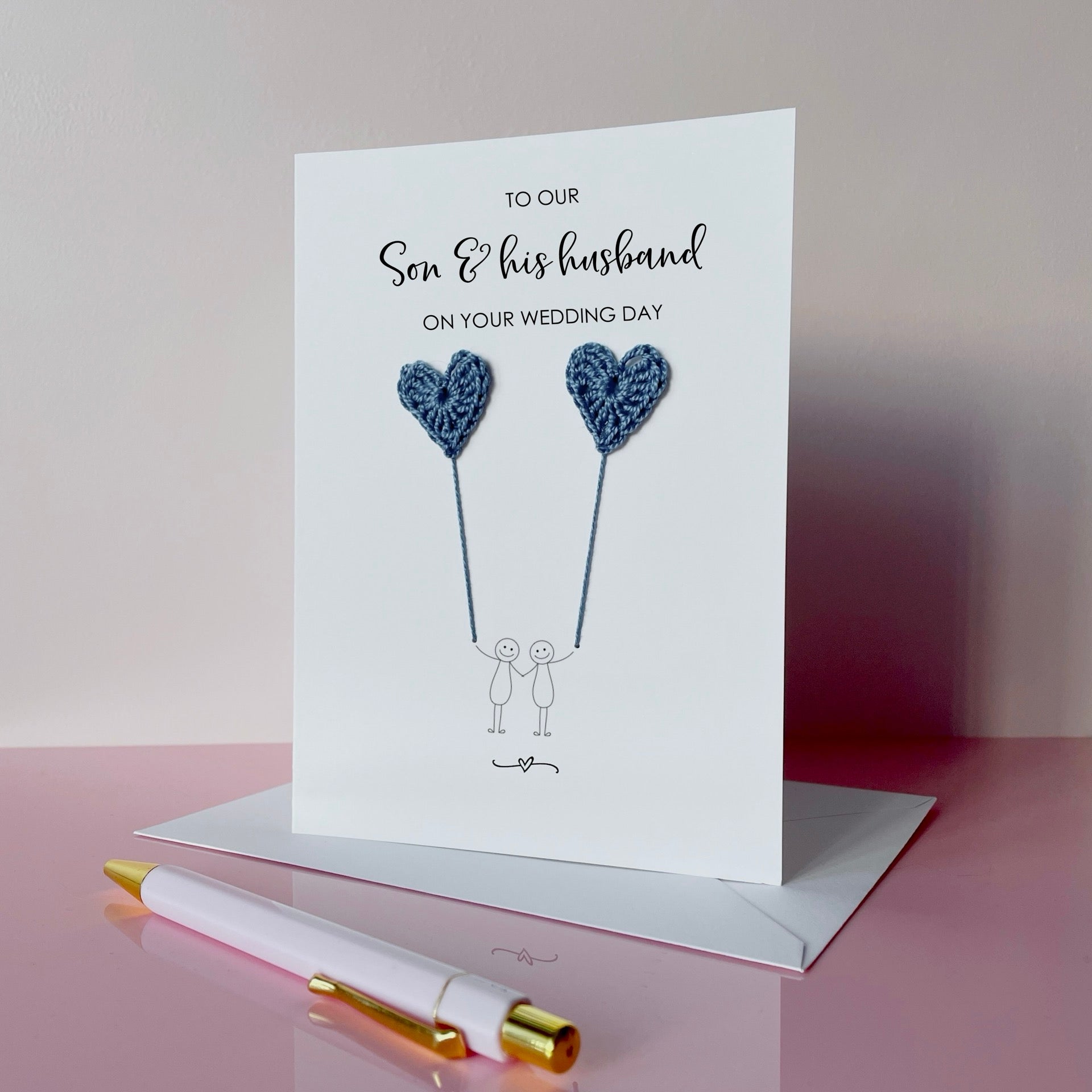 A white greeting card with blue crochet heart balloons. The text at the top of the card reads 'To our Son & his husband on your wedding day' 