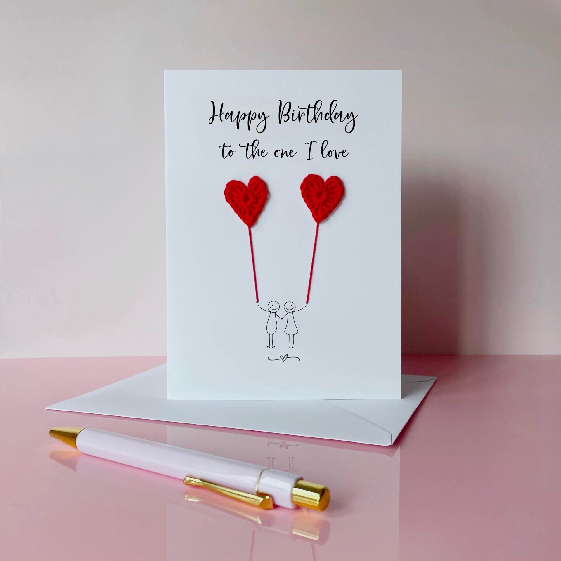 A white birthday card featuring a line drawn couple holding hands and each holding a red crochet heart balloon.  ‘Happy Birthday to the one I love’ is printed at the top of the card in black cursive text.