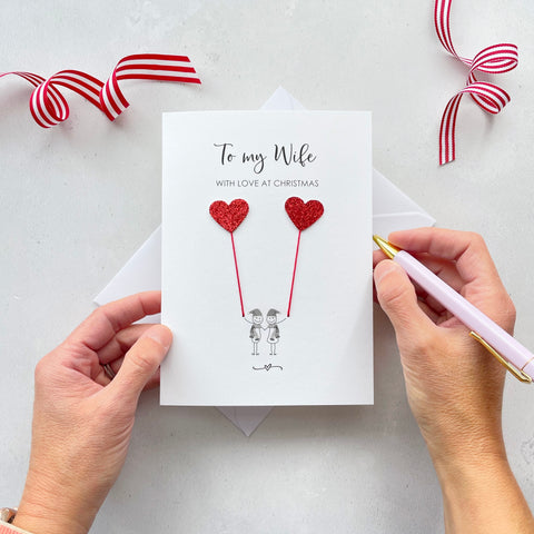 A Christmas card featuring a minimalist design with two figures wearing santa hats holding red glitter heart balloons. The text at the top reads: "To my Wife, With Love at Christmas." The card is placed on a white background with a white envelope partially visible behind it. A hand is seen holding the card, while another hand holds a pink pen. A red and white striped ribbon is positioned in the top corners of the image for decoration.
