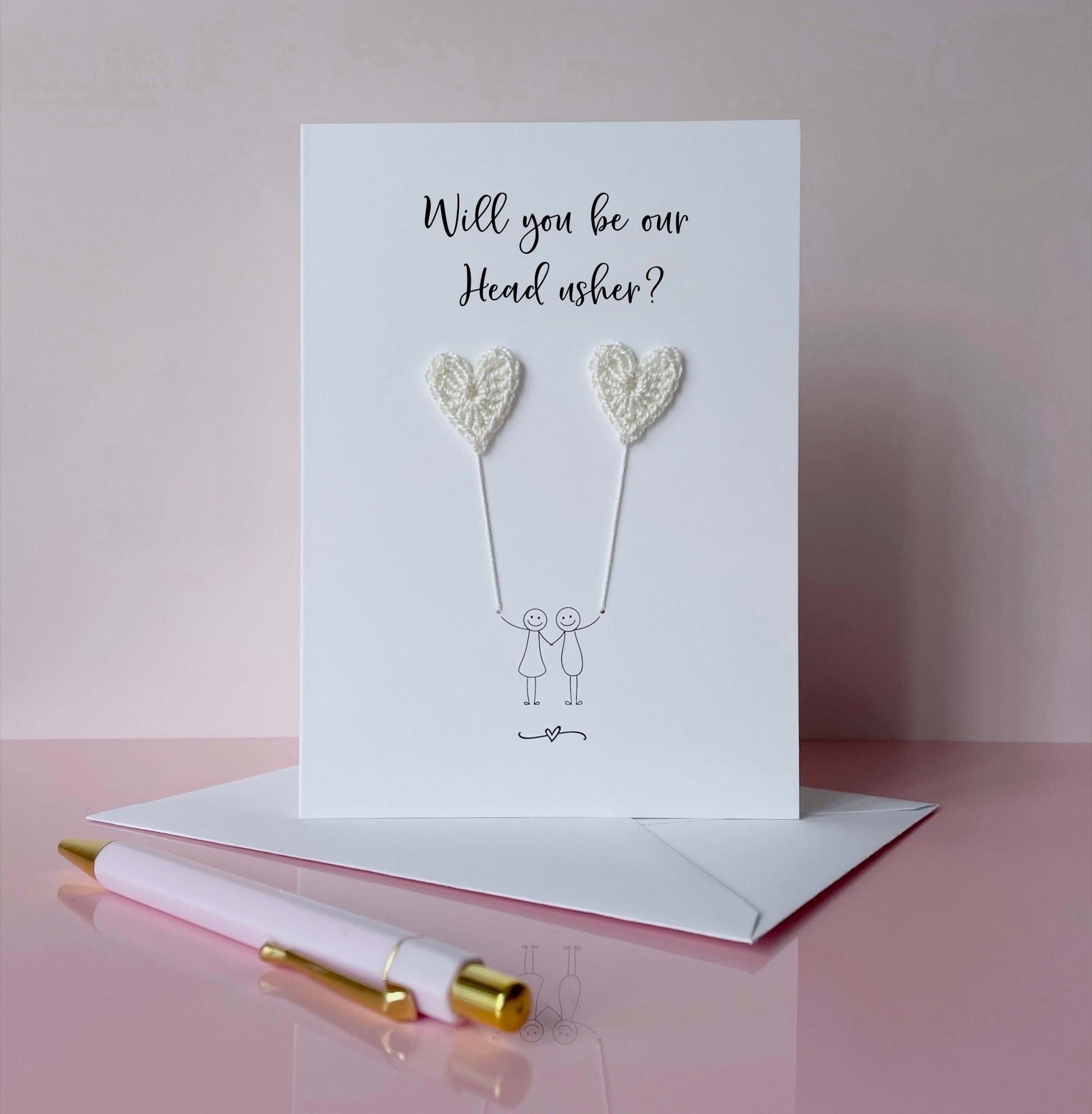 A white card with a line drawn couple printed on it - the couple are each holding a white crochet heart balloon. Will you be our Head usher is printed at the top of the card 