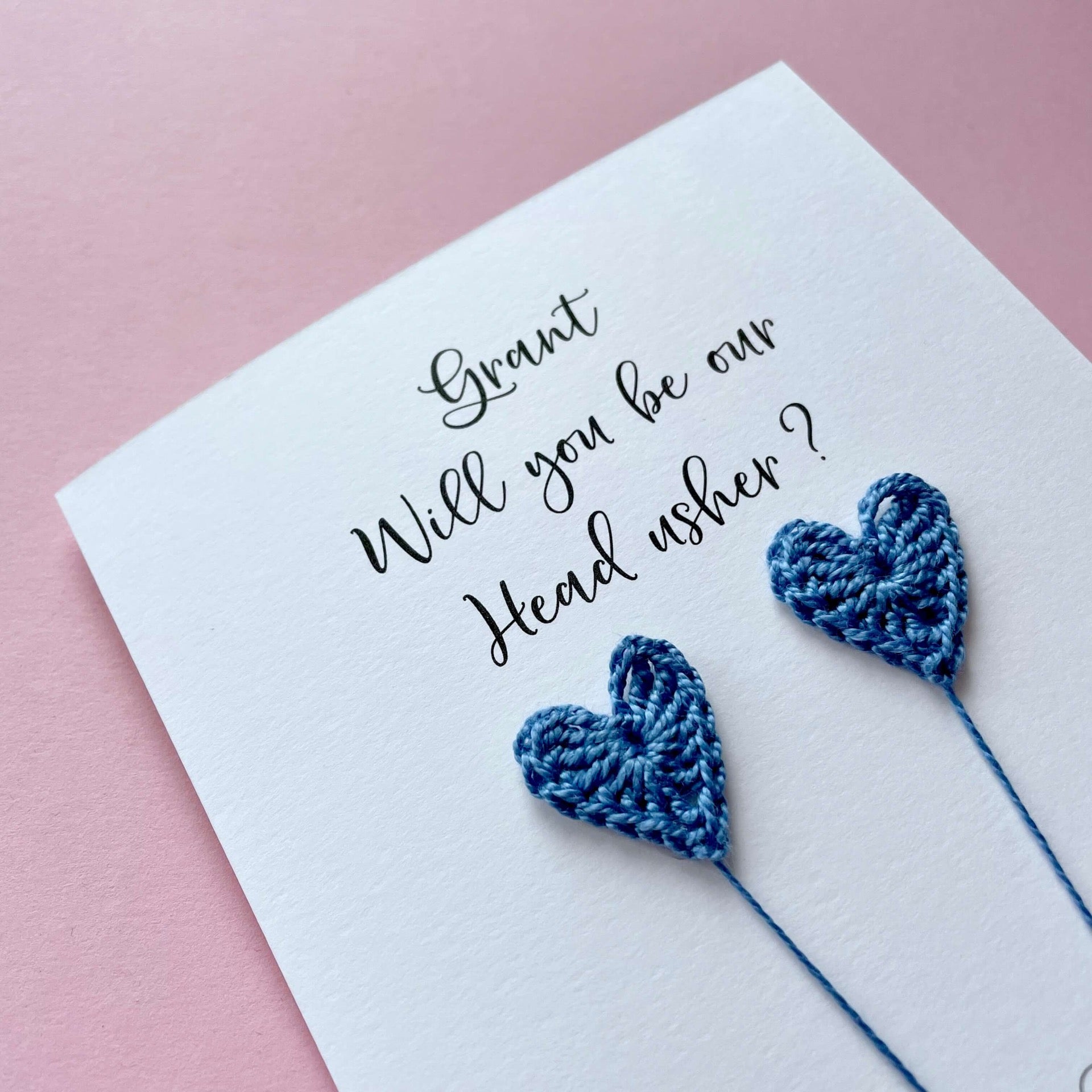 A white card with a line drawn couple printed on it - the couple are each holding a blue crochet heart balloon. Grant, Will you be our Head usher is printed at the top of the card with the option of adding a name, making it the perfect keepsake. 