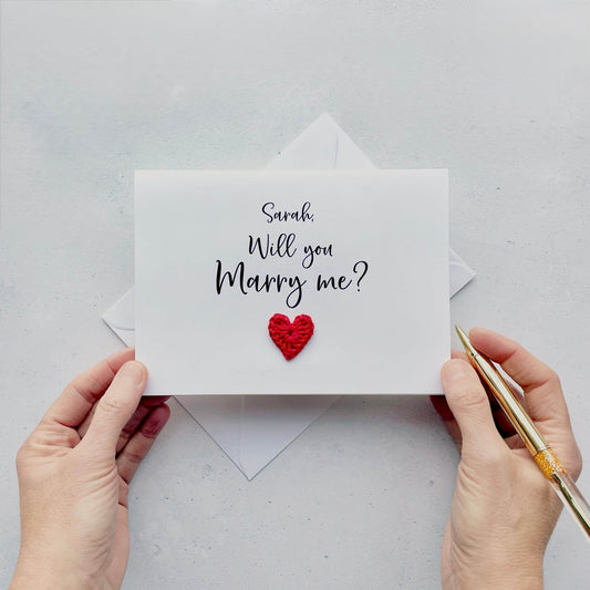 A white card with 'Sarah, Will you marry me?' printed on the front of the card. A small red crochet heart is on the front of the card under this text. 