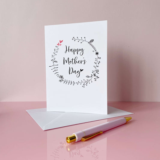 A white Mother’s Day card. Black line drawn foliage is printed on teh card in a circle pattern. The words ‘Happy Mother’s Day’ has been printed inside the circle in black cursive text. 