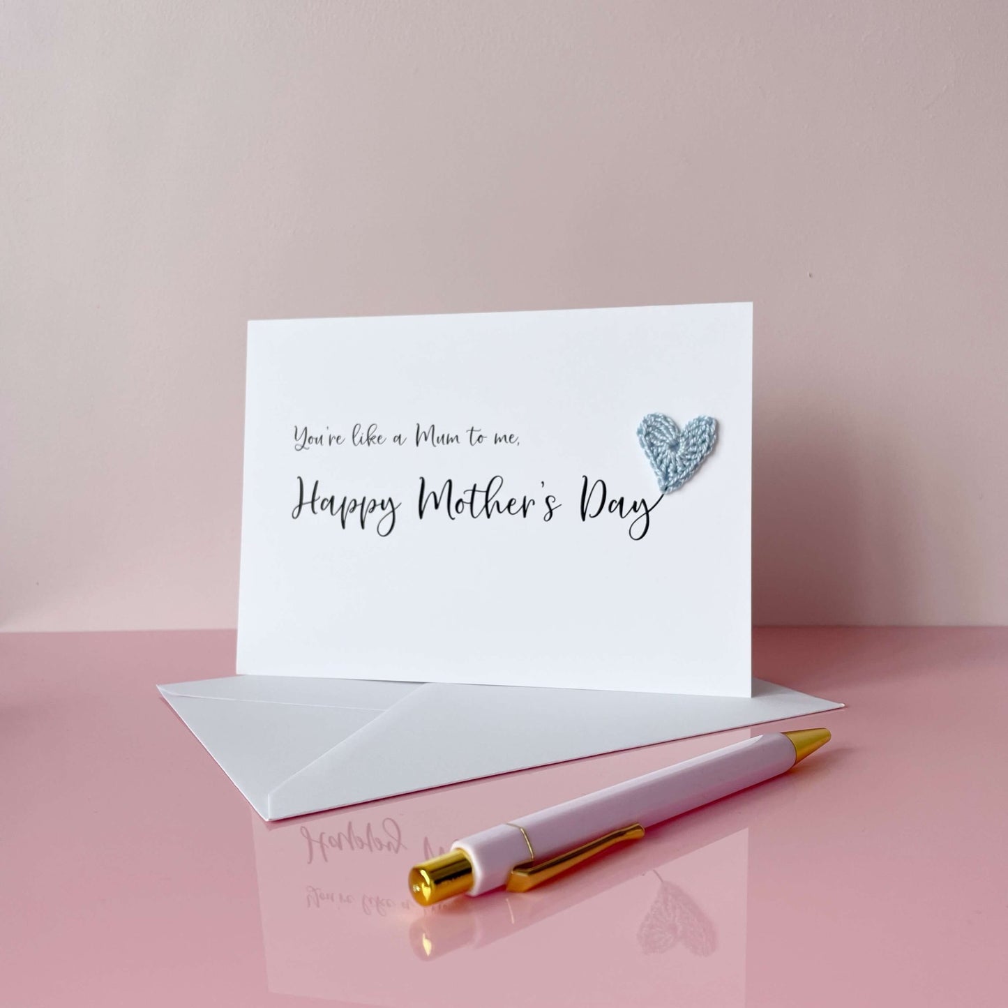 A white Mother’s Day card that has ‘You’re like a Mum to me , Happy Mother’s Day’ printed on the card in black cursive text. A pale blue crochet heart has been attatched to the ‘y’ at the end. 